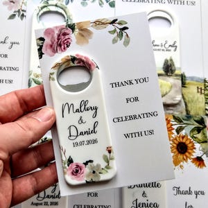 K&ouml;nnte beinhalten: Wei&szlig;er Flaschen&ouml;ffner mit floralen Akzenten und den Namen "Mallory & Daniel" sowie dem Datum "19.07.2026". Eine Karte mit der Aufschrift "Danke, dass Sie mit uns feiern!" in schwarzer Schrift. Hochzeitsgeschenke.
