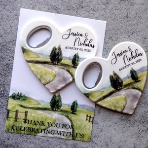 May include: Two heart-shaped bottle openers with a watercolor design of a winding road through a green field. The text "Jessica & Nicholas" and "August 03, 2023" is printed on each opener. The back of the card reads "Thank you for celebrating with us!"