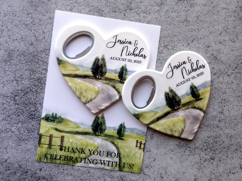 May include: Two heart-shaped bottle openers with a watercolor design of a winding road through a green field. The text "Jessica & Nicholas" and "August 03, 2023" is printed on each opener. The back of the card reads "Thank you for celebrating with us!"