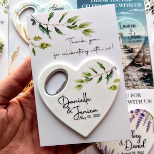 May include: White heart-shaped bottle opener with a green leafy design and the text "Danielle & Jerica May 03, 2025". The bottle opener is attached to a white card with the text "Thank you for celebrating with us!"