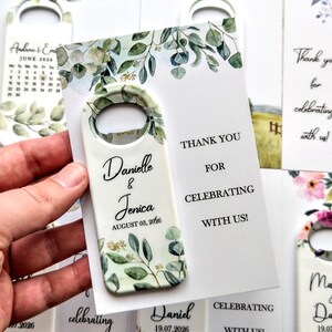 K&ouml;nnte beinhalten: Wei&szlig;er Flaschen&ouml;ffner mit botanischem Design und den Namen "Danielle & Jenica" und dem Datum "03. August 2026". Auf der Karte steht "Danke, dass Sie mit uns feiern!" Hochzeitsgeschenke.