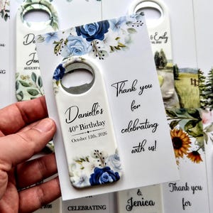 May include: A white bottle opener with a blue floral design and the text "Danielle's 40th Birthday October 12th, 2026." The opener is held next to a card that says "Thank you for celebrating with us!"