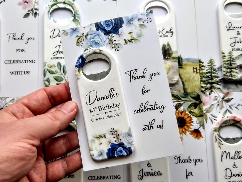 May include: A white bottle opener with a blue floral design and the text "Danielle's 40th Birthday October 12th, 2026." The opener is held next to a card that says "Thank you for celebrating with us!"