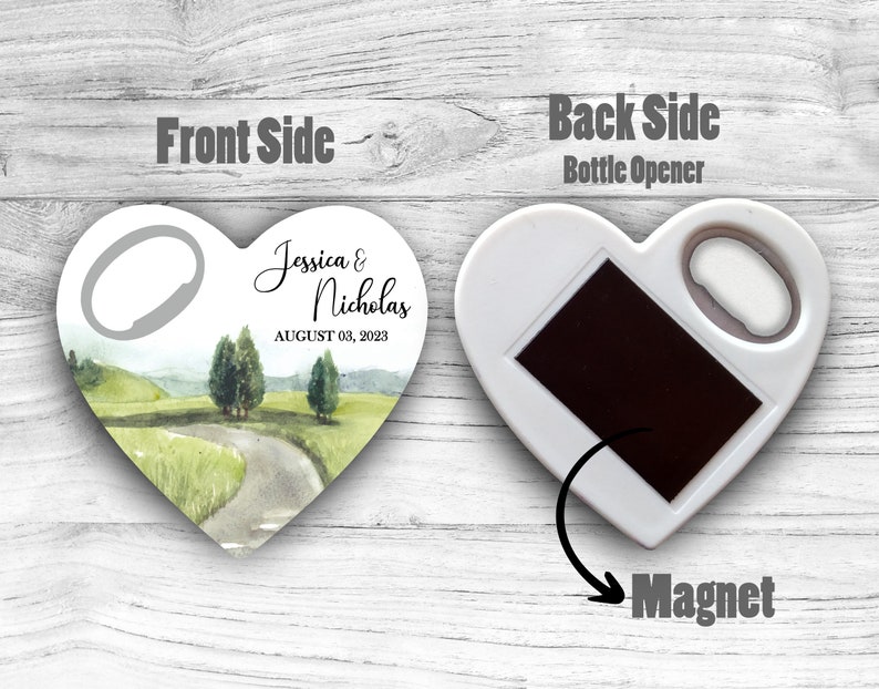 May include: White heart-shaped bottle opener with a magnet on the back. The front side features a watercolor illustration of a winding road through a green landscape with the text "Jessica & Nicholas AUGUST 03, 2023".
