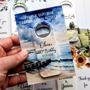 May include: A bottle opener with a beach scene watercolor design. The text reads "Thank you for celebrating with us!" and "Olivia's 60th Birthday 10.09.2026". The opener has a white handle with a hole for hanging.