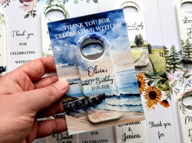May include: A bottle opener with a beach scene watercolor design. The text reads "Thank you for celebrating with us!" and "Olivia's 60th Birthday 10.09.2026". The opener has a white handle with a hole for hanging.