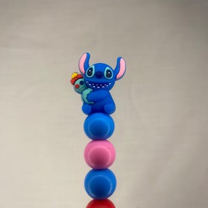 Peut inclure: Une figurine bleue de Stitch tenant un personnage plus petit bleu et rouge, sur une pile de sphères colorées. Les sphères sont bleues, roses, rouges et turquoise. La figurine Stitch a de grandes oreilles et une expression amicale.