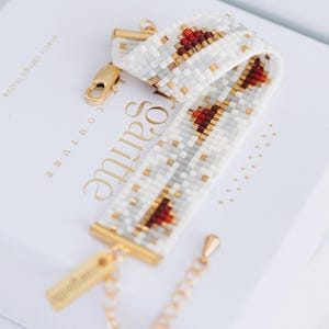 Może przedstawiać: Biała bransoletka z koralików z geometrycznym wzorem w kolorze czerwonym i złotym. Bransoletka jest prezentowana na białym pudełku ze złotymi elementami i złotym łańcuszkiem. Na pudełku widnieje napis "Santie".