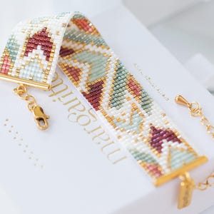 Puede incluir: Una pulsera de cuentas con un patrón geométrico en blanco, burdeos, dorado y turquesa. La pulsera tiene cierres dorados y un extensor de cadena. Se muestra en una caja blanca con el nombre "margari" impreso.