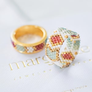 Può includere: Due anelli di perline. Un anello è dorato con un design di perline rosse, bianche e verdi. L'altro anello è bianco con un design di perline rosse, dorate e verdi. Gli anelli sono su una superficie bianca con il testo "marselle JEWELRY".