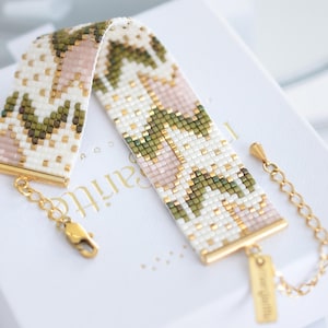 Könnte beinhalten: Ein Perlenarmband mit einem floralen Muster in Weiß, Rosa, Grün und Gold. Das Armband hat goldfarbene Metallakzente und einen Kettenverschluss. Es wird auf einer weißen Schachtel präsentiert.