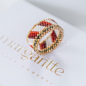 May include: A beaded ring with a geometric pattern in red, white, and green. The ring has a gold-colored band and is set on a white surface with the text "margartte jewelry couture".
