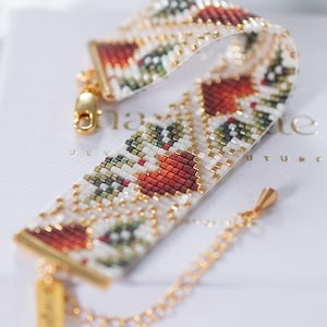 Può includere: Un braccialetto di perline con chiusura e catena dorate. Il braccialetto presenta un motivo geometrico di perline rosse, verdi e bianche. Una piccola etichetta dorata pende dal braccialetto. Il braccialetto è esposto su una superficie bianca.