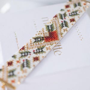May include: A beaded bracelet with a white background. The bracelet features a repeating pattern of red, green, and gold beads. The bracelet is resting on a white box with gold lettering. The bracelet is approximately 18 cm long.