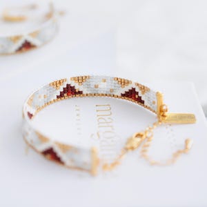 May include: A beaded bracelet with a geometric pattern in white, gold, and burgundy. The bracelet is on a white box with the word "margaret" visible. The bracelet has a gold clasp and chain.