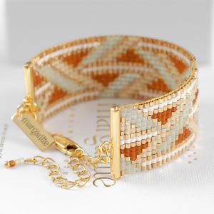 以下が含まれることがあります： オレンジ、白、ペールグリーンの幾何学模様が施された、幅広のビーズブレスレット。ブレスレットにはゴールドカラーのメタルアクセントと留め金が付いています。「margartitte」のブランド名が小さなタグに表示されています。
