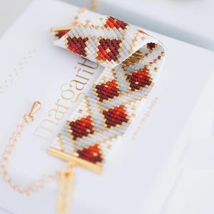 May include: A beaded bracelet with a geometric pattern of red and white beads, accented with gold-colored metal. The bracelet is resting on a white box with the word "margarita" in gold lettering. The bracelet is approximately 7 inches long.