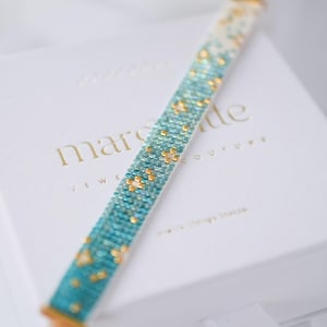 May include: A turquoise and gold beaded bracelet, displayed on a white box embossed with the words "marcade JEWELRY COUTURE". The bracelet has a gradient effect, with a pattern of small, square beads. The box also has the words "more things inside".