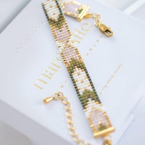 May include: A beaded bracelet with a gold clasp and extender chain. The bracelet features a pattern of white, pink, green, and gold beads. The bracelet is resting on a white box with gold lettering.