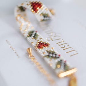 Può includere: Un braccialetto di perline con base bianca, con accenti rossi e verdi in un motivo ad albero di Natale. Il braccialetto ha una chiusura e una catena color oro, su sfondo bianco.