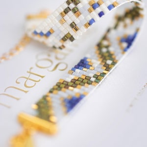 May include: Close-up of a beaded bracelet with a white background. The bracelet features a woven pattern of white, gold, blue, and green beads. The clasp and chain are gold. The bracelet is a piece of jewelry.