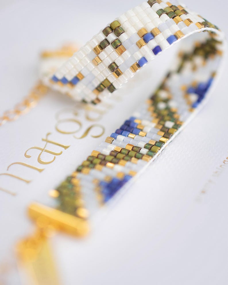 May include: Close-up of a beaded bracelet with a white background. The bracelet features a woven pattern of white, gold, blue, and green beads. The clasp and chain are gold. The bracelet is a piece of jewelry.