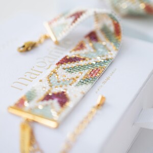 May include: A beaded bracelet with a geometric pattern in white, gold, green, and burgundy. The bracelet has gold-colored clasps and a small tag. The bracelet is resting on a white box with gold lettering.