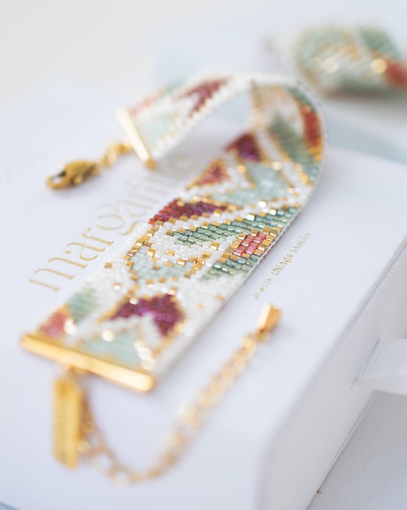 May include: A beaded bracelet with a geometric pattern in white, gold, green, and burgundy. The bracelet has gold-colored clasps and a small tag. The bracelet is resting on a white box with gold lettering.