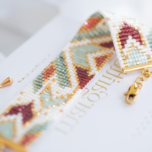 May include: A beaded bracelet with a geometric pattern in white, red, green, and gold. The bracelet is made of small beads and has a gold clasp. The design features a repeating pattern of shapes and colors.