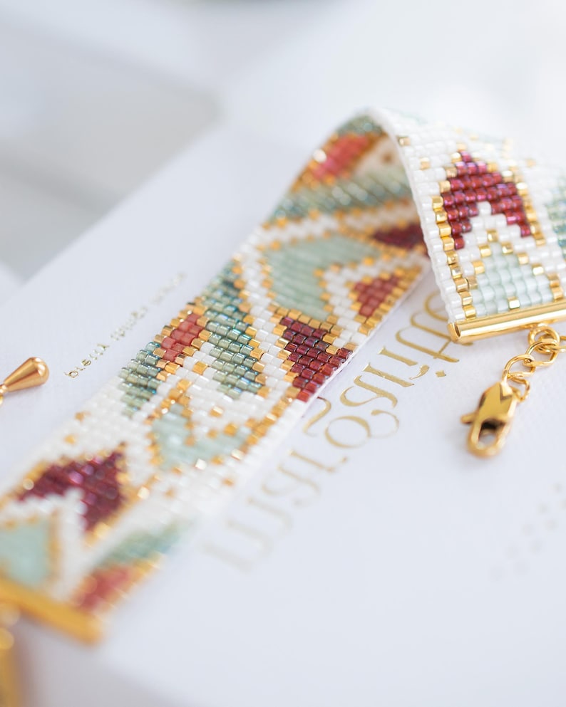 May include: A beaded bracelet with a geometric pattern in white, red, green, and gold. The bracelet is made of small beads and has a gold clasp. The design features a repeating pattern of shapes and colors.