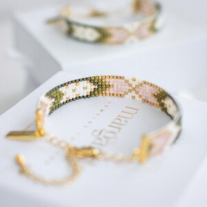 Op de afbeelding: Twee armbanden met kralen met een geometrisch patroon in wit, roze, groen en goud. De armbanden liggen op een wit oppervlak. E&eacute;n armband is scherp, met een gouden sluiting en ketting.