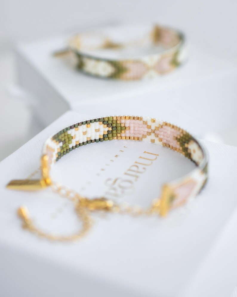 Op de afbeelding: Twee armbanden met kralen met een geometrisch patroon in wit, roze, groen en goud. De armbanden liggen op een wit oppervlak. E&eacute;n armband is scherp, met een gouden sluiting en ketting.