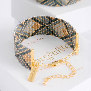 Op de afbeelding: Een kralenarmband met een geometrisch patroon in blauw, grijs en goud. De armband heeft goudkleurige metalen accenten en een bijpassende ketting. Het woord "margritte" is zichtbaar op de doos.