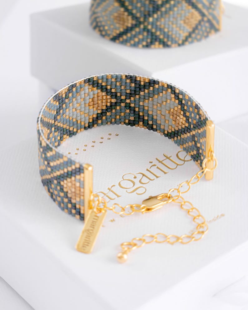Op de afbeelding: Een kralenarmband met een geometrisch patroon in blauw, grijs en goud. De armband heeft goudkleurige metalen accenten en een bijpassende ketting. Het woord "margritte" is zichtbaar op de doos.