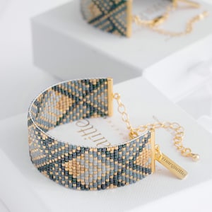 Op de afbeelding: Een kralenarmband met een geometrisch patroon in grijstinten, goud en zwart. De armband heeft goudkleurige metalen accenten en een klein label met de tekst "margartie". Een andere armband is zichtbaar op de achtergrond.