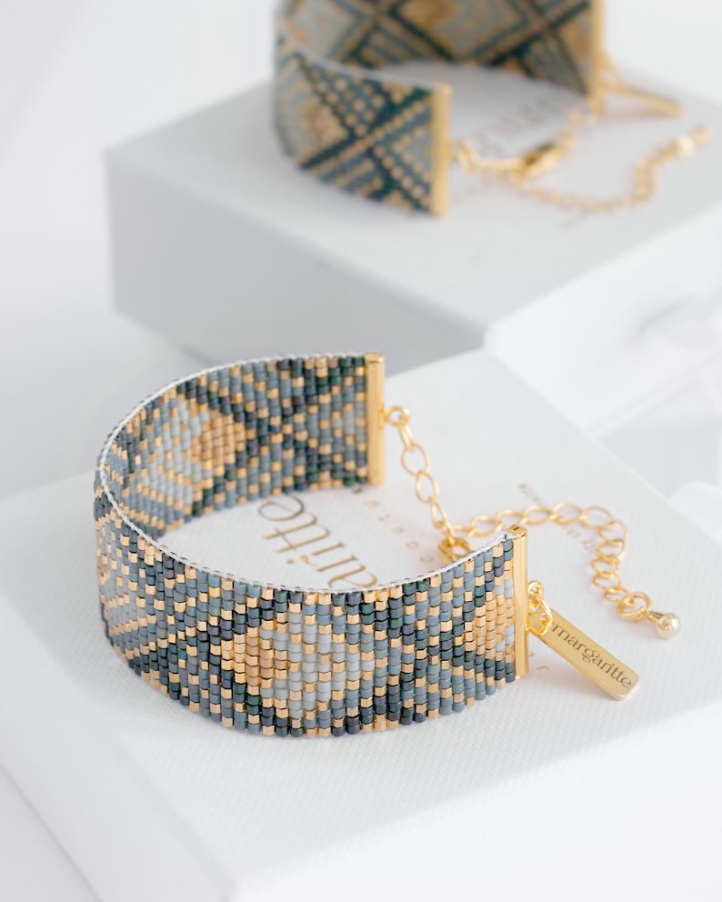 Op de afbeelding: Een kralenarmband met een geometrisch patroon in grijstinten, goud en zwart. De armband heeft goudkleurige metalen accenten en een klein label met de tekst "margartie". Een andere armband is zichtbaar op de achtergrond.