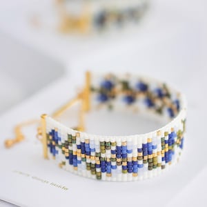 May include: A white beaded bracelet with a floral pattern in blue, green, and gold. The bracelet has gold-colored metal accents and a delicate chain closure. The text "jewelry things inside" is visible on the surface.