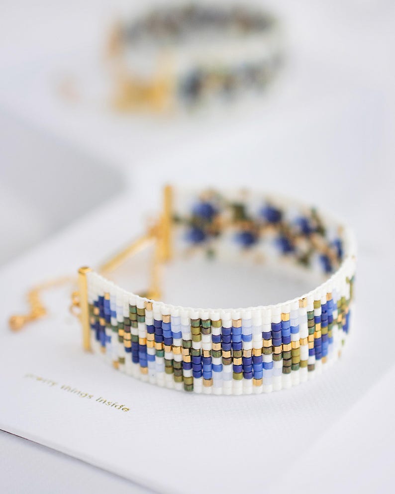 May include: A white beaded bracelet with a floral pattern in blue, green, and gold. The bracelet has gold-colored metal accents and a delicate chain closure. The text "jewelry things inside" is visible on the surface.