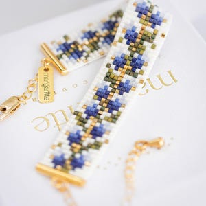 May include: A beaded bracelet with a floral pattern in blue, green, and gold on a white background. The bracelet has gold-colored metal accents and a small tag that says "margartte". The bracelet is on a white surface.
