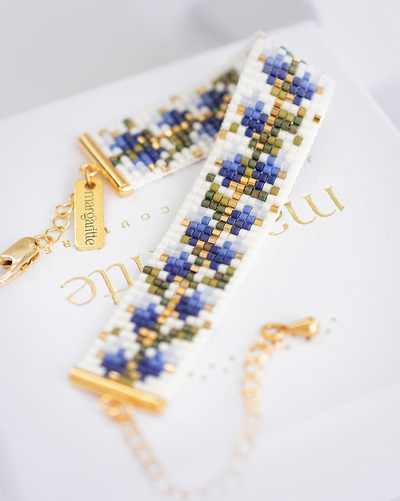 May include: A beaded bracelet with a floral pattern in blue, green, and gold on a white background. The bracelet has gold-colored metal accents and a small tag that says "margartte". The bracelet is on a white surface.