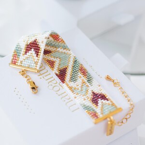 May include: A beaded bracelet with a geometric pattern in white, burgundy, gold, and teal. The bracelet has gold-colored clasps and a chain extender. The bracelet is displayed on a white box with the name "margari" printed on it.