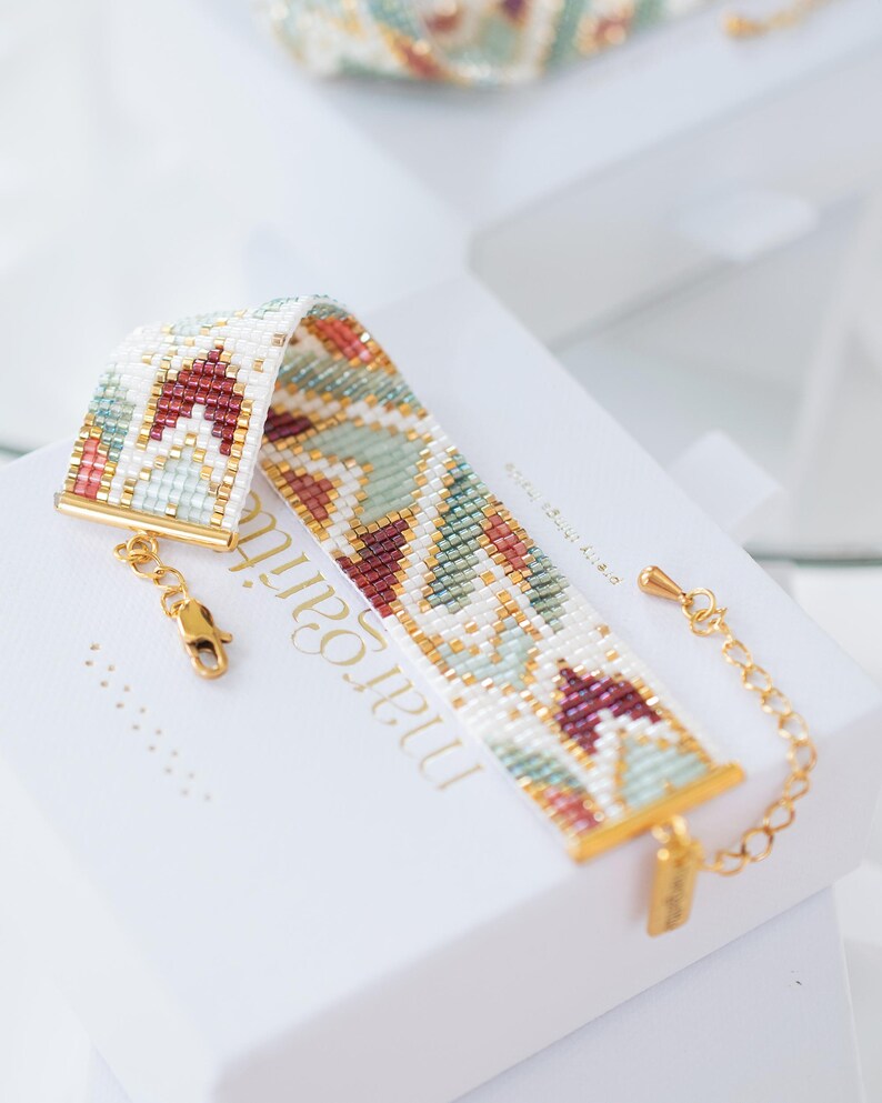 May include: A beaded bracelet with a geometric pattern in white, burgundy, gold, and teal. The bracelet has gold-colored clasps and a chain extender. The bracelet is displayed on a white box with the name "margari" printed on it.