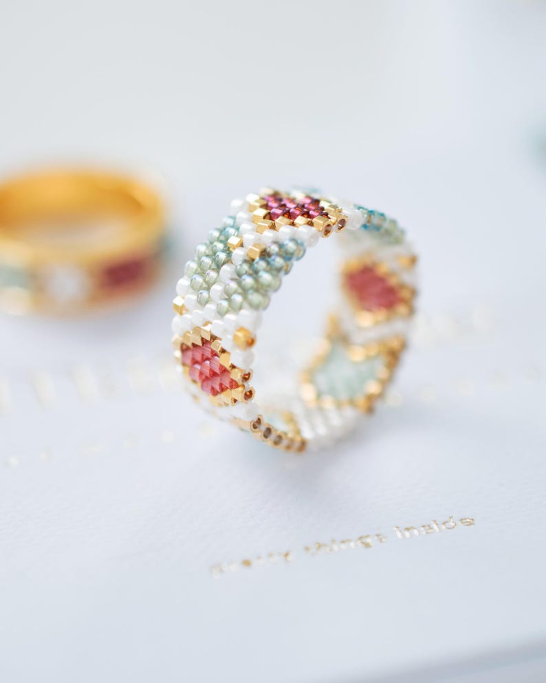May include: A beaded ring with a geometric pattern in white, gold, pink, and teal. The ring is in focus, with a blurred gold ring in the background. The ring's design features small beads arranged in a repeating pattern.