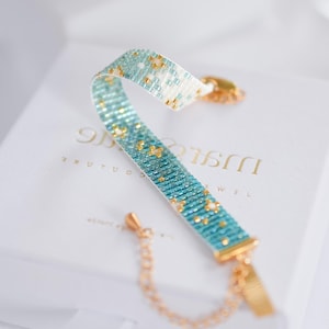 May include: A beaded bracelet with a turquoise and gold pattern. The bracelet is made of small, colorful beads and has a gold-colored clasp and chain. The bracelet is resting on a white box.