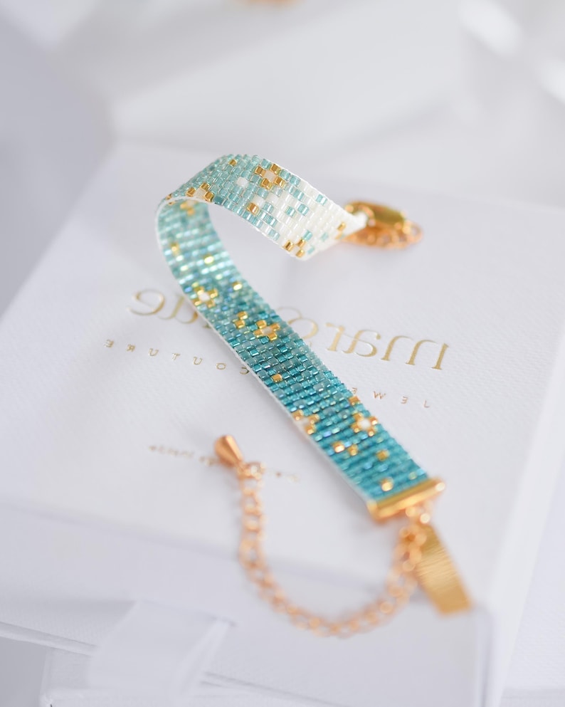 May include: A beaded bracelet with a turquoise and gold pattern. The bracelet is made of small, colorful beads and has a gold-colored clasp and chain. The bracelet is resting on a white box.