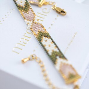 Op de afbeelding: Een kralenarmband met een geometrisch patroon in groen, wit, goud en roze. De armband heeft een gouden sluiting en ketting. De armband ligt op een witte doos met gouden tekst.