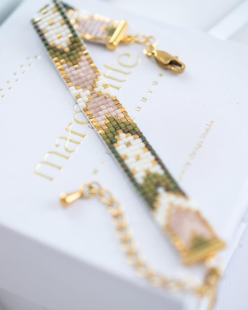 Op de afbeelding: Een kralenarmband met een geometrisch patroon in groen, wit, goud en roze. De armband heeft een gouden sluiting en ketting. De armband ligt op een witte doos met gouden tekst.