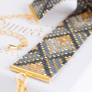 Op de afbeelding: Een kralenarmband met een geometrisch patroon in goud, blauw en grijs. De armband heeft goudkleurige metalen accenten en een delicate ketting. Het ontwerp is ingewikkeld en gedetailleerd, wat het vakmanschap van de sieraden laat zien.