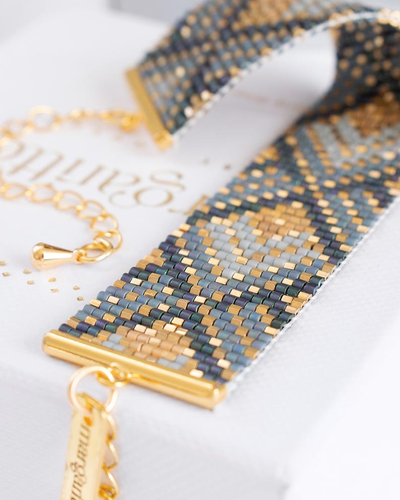Op de afbeelding: Een kralenarmband met een geometrisch patroon in goud, blauw en grijs. De armband heeft goudkleurige metalen accenten en een delicate ketting. Het ontwerp is ingewikkeld en gedetailleerd, wat het vakmanschap van de sieraden laat zien.