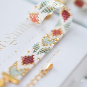 May include: A beaded bracelet with a white background, featuring a repeating pattern of red, green, and gold beads. The bracelet has a gold clasp and chain. The bracelet is on top of a white box.